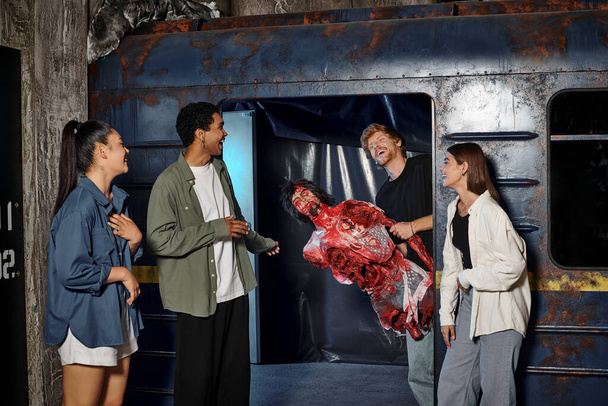 joyeux jeune homme tenant mannequin effrayant et effrayant amis multiculturels dans la salle d'évasion - Photo, image