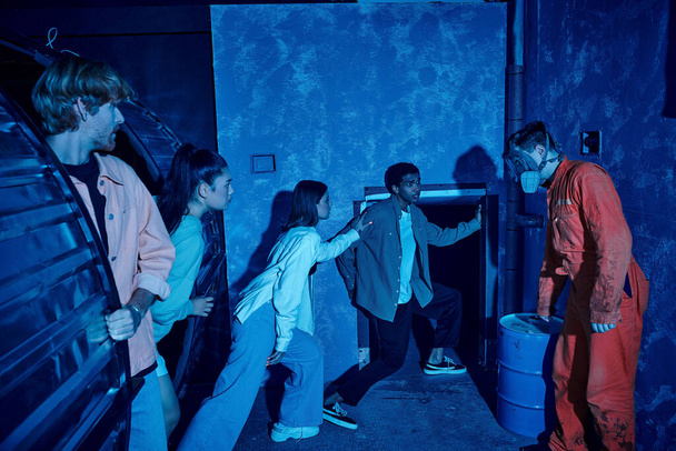 Grupo multicultural de amigos escapando del hombre con máscara de gas durante la aventura en la sala de misiones - Foto, imagen