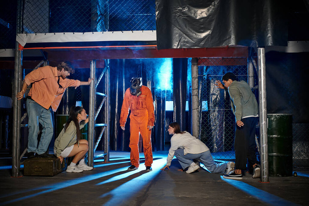 personne bizarre en costume de ppe orange et masque à gaz près de jeunes amis interraciaux dans la salle d'évasion - Photo, image
