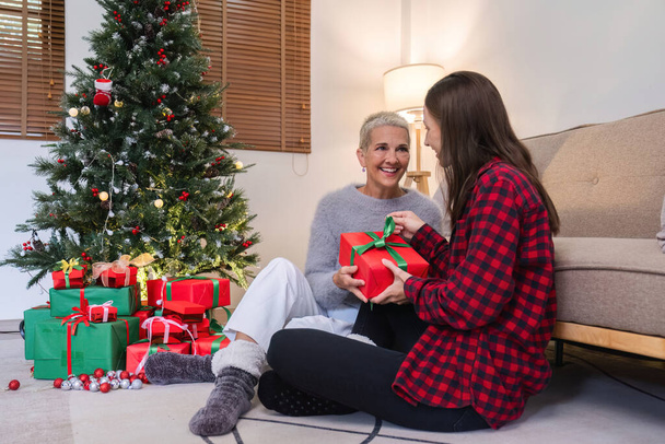 Милая молодая женщина держит подарочную коробку, чтобы удивить свою мать на Рождество. 60-летняя женщина на пенсии счастливая дочь на Рождество в гостиной с красной подарочной коробкой. - Фото, изображение