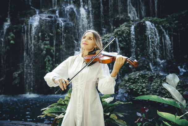Beautiful Caucasian woman playing violin near waterfall. Music and art concept. Pretty female wearing white dress in nature. Water splash. Banyu Wana Amertha waterfall Wanagiri, Bali, Indonesia. - Photo, Image