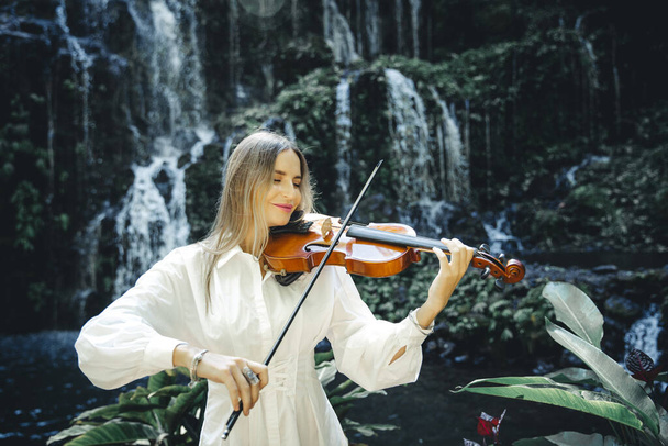 Affascinante donna caucasica che suona il violino a occhi chiusi vicino alla cascata. Concetto musicale e artistico. Donna con lunghi capelli biondi vestita di bianco in natura. Un goccio d'acqua. Cascata di Banyu Wana Amertha - Foto, immagini