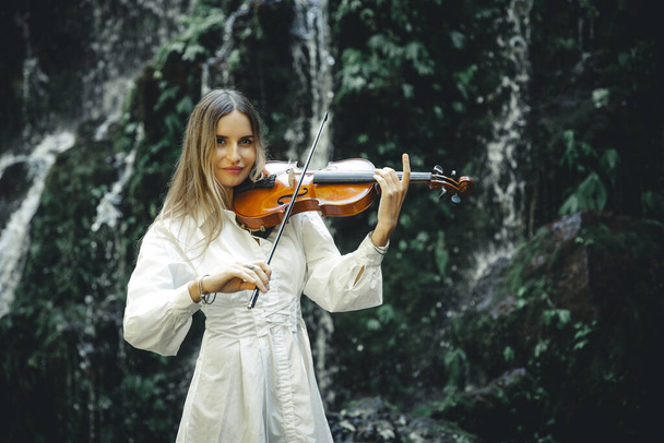 Красива кавказька жінка грає на скрипці біля водоспаду. Концепція музики та мистецтва. Жінка в білій сукні в природі. Сплеск води. Тропічний ліс. Балі - водоспад Амерта, Балі. Копіювати простір. - Фото, зображення