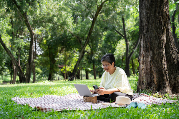 Mulher sênior asiática feliz usando laptop para trabalhar on-line fora do escritório. Reforma idosa usando laptop para aprender novas habilidades e freelance trabalhando no parque, estilo de vida sênior com tecnologia - Foto, Imagem