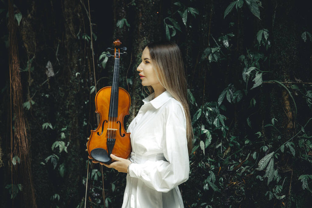 Portrait d'une charmante femme caucasienne tenant un violon dans une forêt tropicale. Vue de profil de visage de femme. Concept musical et artistique. Femme en robe blanche. Fond de feuilles tropicales vertes luxuriantes. Bali - Photo, image