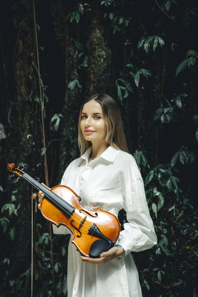 Portrait of beautiful Caucasian woman holding violin in tropical forest. Music and art concept. Female wearing white dress. Background of lush green tropical leaves. Bali. Copy space. Vertical layout. - Photo, Image