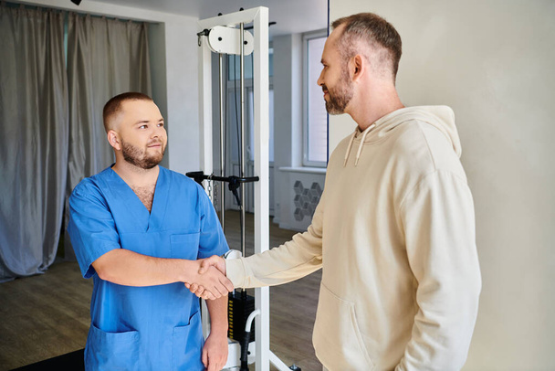 vousatý muž třesoucí se ruce s úsměvem lékař v modré uniformě v rehabilitační kinezio centrum - Fotografie, Obrázek