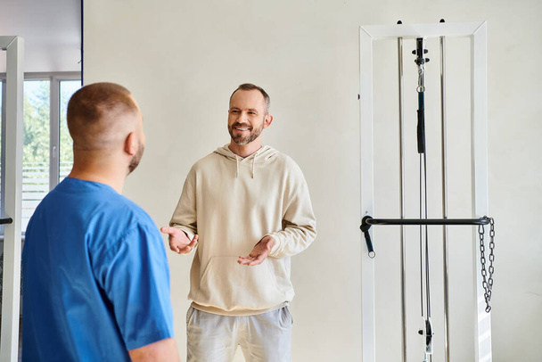 positive man talking to doctor in blue uniform during appointment in rehabilitation kinesio center - Photo, Image