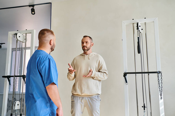 paziente di sesso maschile discutere piano di trattamento con il medico in uniforme blu nel centro di recupero kinesio - Foto, immagini