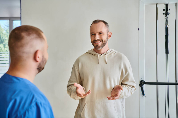 щасливий бородатий чоловік жестикулює і розмовляє з молодим фахівцем в реабілітаційному центрі кінезіотерапії - Фото, зображення