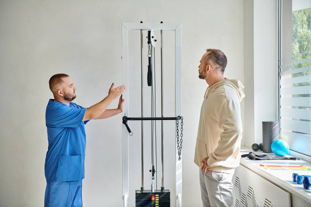rehabilitolog v modré uniformě ukazující cvičební pomůcky svému pacientovi v kineziologickém centru - Fotografie, Obrázek