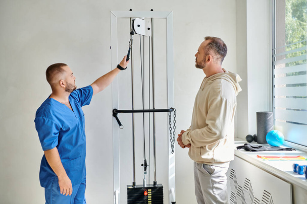 professional rehabilitologist in blue uniform showing training machine to man in kinesiology center - Fotografie, Obrázek