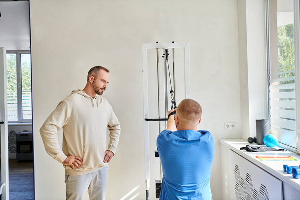 Mann schaut auf Erholungsexperten in blauer Uniform beim Training an Trainingsgerät im Kinesiologischen Zentrum - Foto, Bild