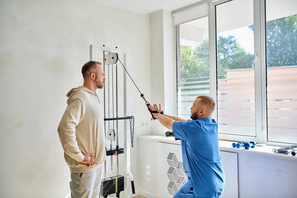 fiatal rehabilitológus, aki megmutatja a férfi betegnek, hogyan kell edzeni a gyakorló gépen a kinesio központban - Fotó, kép