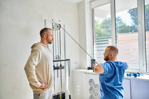 professionele arts tonen mannelijke patiënt hoe uit te werken op fitnessapparaat in kinesio centrum - Foto, afbeelding