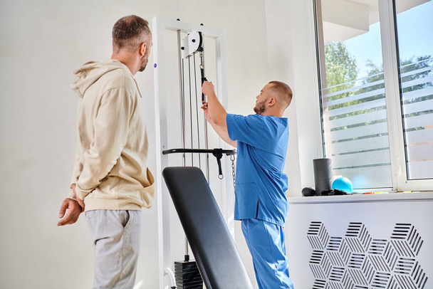 специалист по восстановлению в синей форме, показывающий тренировочное оборудование пациенту мужского пола в центре Кинезио - Фото, изображение