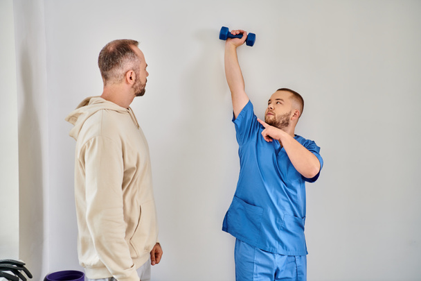 skilled rehabilitologist showing exercise with dumbbell to his patient in recovery kinesio center - Photo, Image