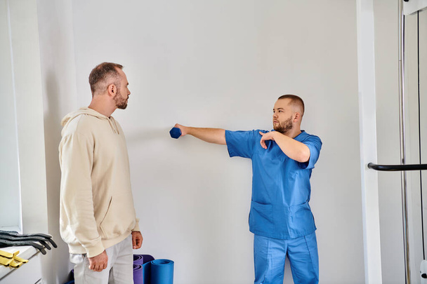 опытный реабилитолог показывает упражнения с гантелями человеку в реабилитационном центре Кинезио - Фото, изображение