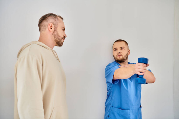 Junge und erfahrene Ärztin zeigt Mann im Rehabilitationskinesio-Zentrum, wie man mit Hantel trainiert - Foto, Bild