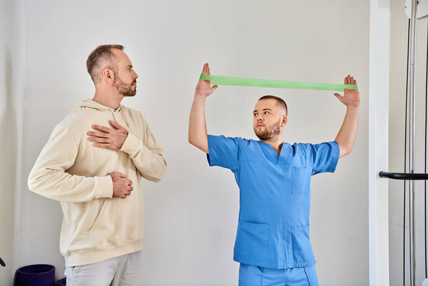 Rehabilitologe zeigt dem Mann mit verletzter Schulter Übung mit Widerstandsband, Kinesiozentrum - Foto, Bild