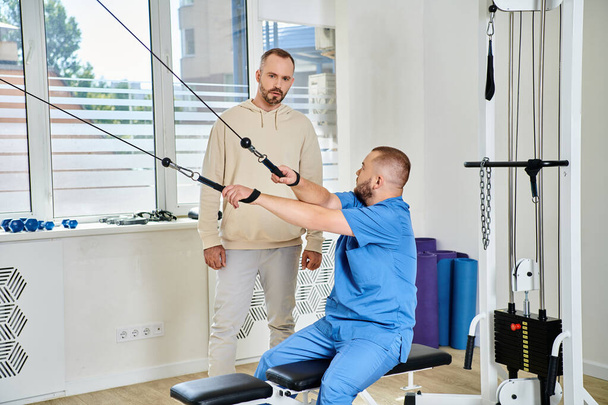 Rehabilitologe in blauer Uniform zeigt dem Mann im Kinesio-Zentrum Übungen an einem Trainingsgerät - Foto, Bild