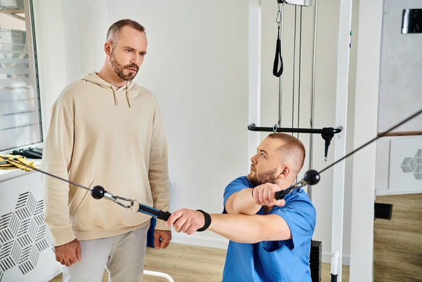 physiotherapist in blue uniform showing exercise on training machine to man in kinesio center - Photo, Image