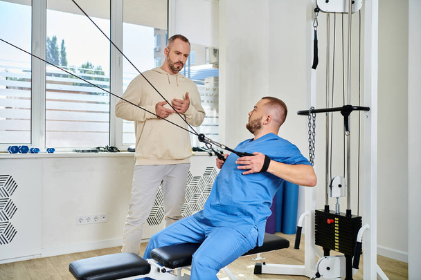 skilled rehabilitation specialist showing exercise on training machine to man in kinesiology center - Photo, Image