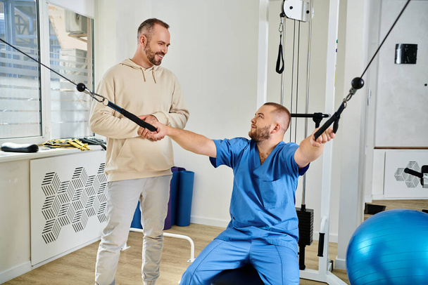 arts in blauw uniform tonen oefening op trainingsmachine naar lachende man in kinesiologie centrum - Foto, afbeelding