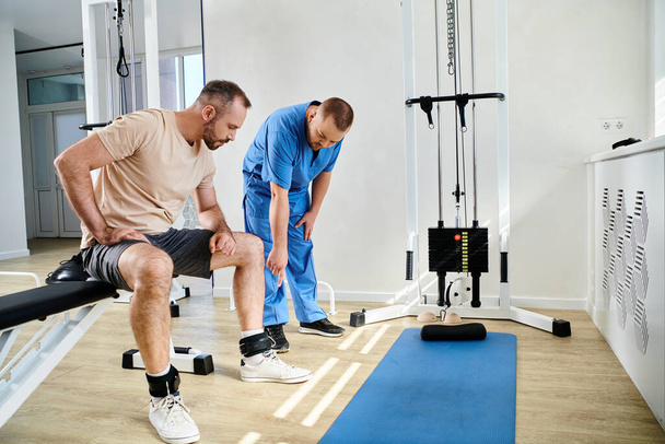 mladý lékař ukazuje na pásy nohou muže sedícího v blízkosti cvičebního stroje v tělocvičně kinezio centra - Fotografie, Obrázek