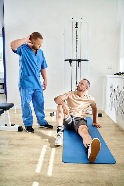 revalidoloog in gesprek met man zittend op fitnessman in de buurt van trainingsapparaat in kinesio centrum - Foto, afbeelding