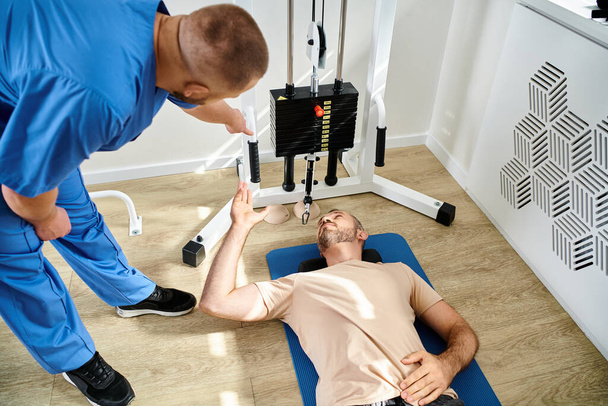 vista ad alto angolo dell'istruttore che assiste il suo paziente sdraiato sul fitness man vicino all'attrezzo da ginnastica - Foto, immagini
