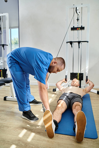 Arzt in blauer Uniform hilft Mann in Sportbekleidung auf Fitnessmatte in der Nähe von Trainingsgerät - Foto, Bild