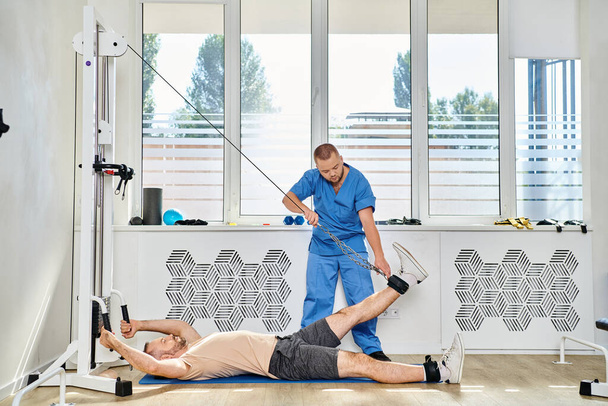 habilidoso rehabilitólogo que ayuda al hombre a ejercitarse en la máquina de ejercicio en el gimnasio del centro kinesio - Foto, Imagen