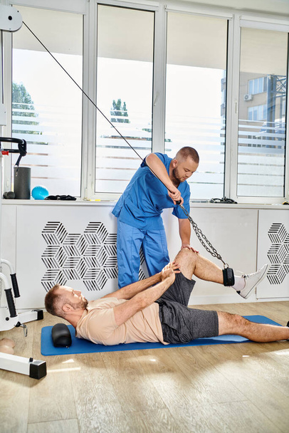 joven especialista en recuperación que ayuda al hombre a ejercitarse en la máquina de ejercicio en el gimnasio del centro kinesio - Foto, Imagen