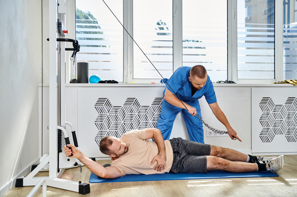 молодой врач в синей форме помогает мужчине тренироваться на тренажере в тренажерном зале кинезио-центра - Фото, изображение
