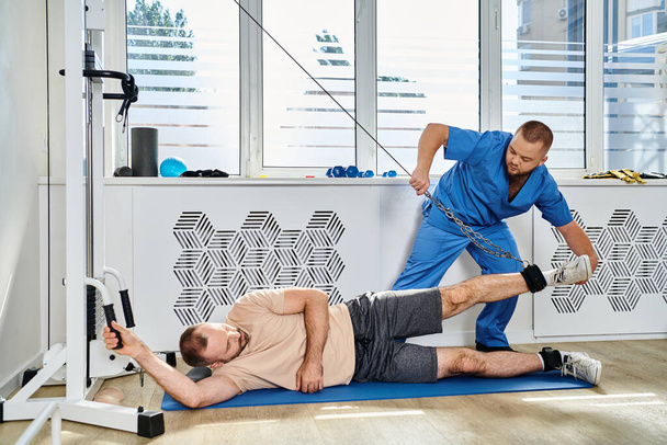 profesionální asistent pomáhající muži trénovat na moderním cvičebním stroji v tělocvičně kinezio centra - Fotografie, Obrázek