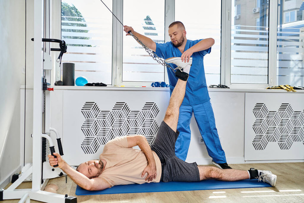 Junge Rehabilitologin hilft Mann beim Training auf modernem Trainingsgerät in Turnhalle des Kinesio-Zentrums - Foto, Bild
