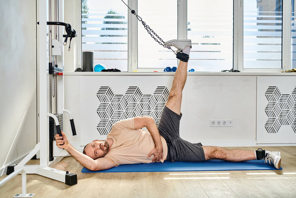 man in sportswear lying down on fitness mat and training leg on exercise machine in kinesio center - Photo, Image