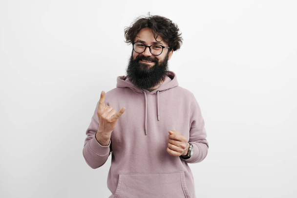 Веселий бородатий чоловік в модному светрі і окулярах, блимає камінь на знак руки з грайливою посмішкою на чистому білому тлі - Фото, зображення
