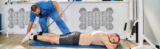 jonge rehabilitoloog helpen man training op oefening machine in moderne kinesio centrum, banner - Foto, afbeelding