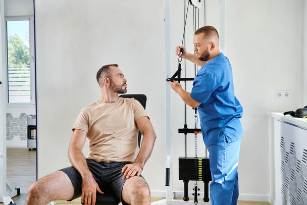 Genesungsspezialist erklärt männlichen Patienten, wie man an einem Trainingsgerät im Kinesio-Zentrum trainiert - Foto, Bild