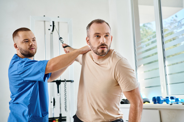 young physician in blue uniform helping man working out on training machine in kinesiology center - Photo, Image