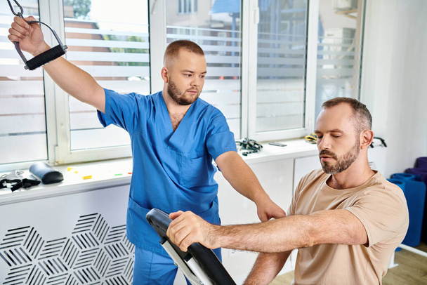 bearded man in sportswear training on exercise machine near young physician in kinesio center - Photo, Image