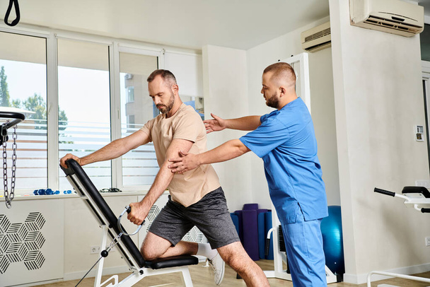 instructor profesional que apoya al hombre en ropa deportiva durante el entrenamiento de recuperación en la máquina de ejercicio - Foto, imagen