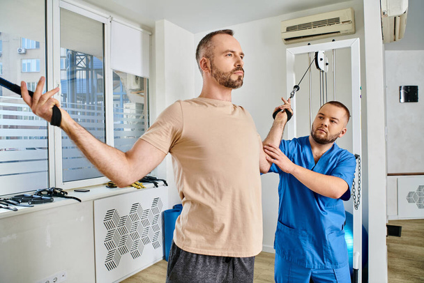 Facharzt hilft Mann beim Sportbekleidungstraining an Trainingsgerät im modernen Kinesio-Zentrum - Foto, Bild