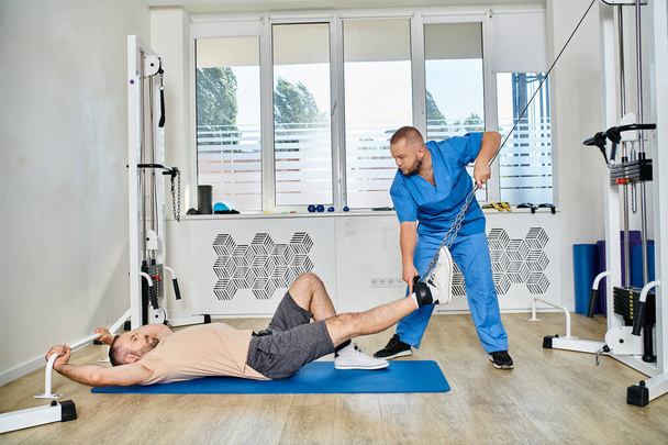 mladý lékař v modré uniformě pomáhá muž školení na cvičební stroj v tělocvičně kinesio centra - Fotografie, Obrázek