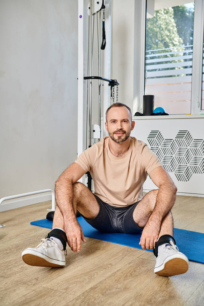 smiling bearded man in sportswear sitting on fitness mat near exercise machine in kinesio center - Photo, Image