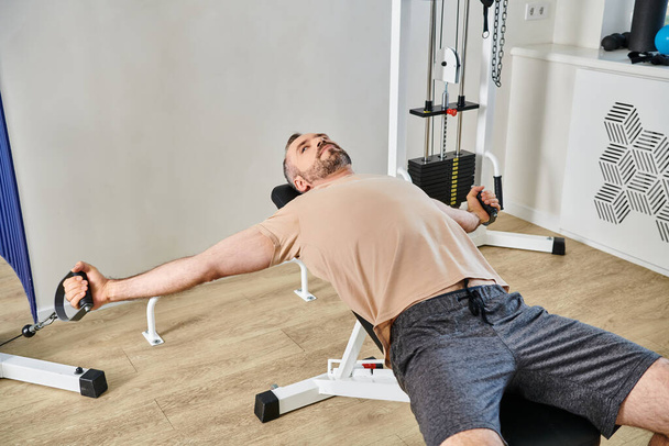 bebaarde man in sportkleding uit te werken op de oefening machine in kinesio centrum, herstel sessie - Foto, afbeelding