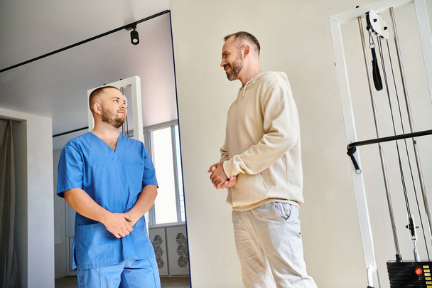 досвідчений реабілітолог у блакитній формі розмовляє з пацієнтом чоловічої статі в сучасному центрі кінезіо - Фото, зображення