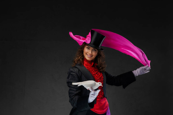 Una mujer ilusionista elegante, vestida con un disfraz de magos y un sombrero de cilindro negro, realiza trucos fascinantes con una elegante paloma blanca y un pañuelo de seda violeta contra un telón de fondo oscuro - Foto, Imagen